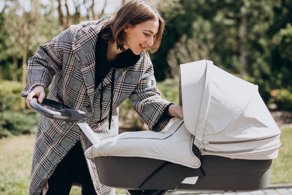 Uśmiechnięta mama zagląda do gondoli podczas spaceru w ogrodzie, prezentując komfortowe polskie wózki dziecięce